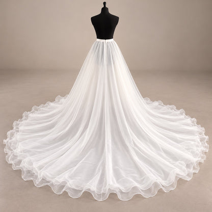 A white organza wedding dress displayed on a black mannequin torso against a plain beige studio background. The dress has a fitted bodice and a very full, flowing skirt with a long train spread wide across the floor.