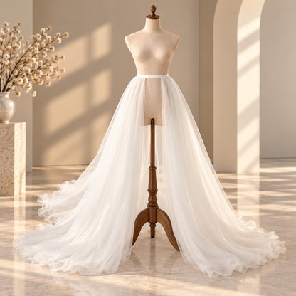 A white tulle wedding dress displayed on a beige mannequin torso with a wooden stand, set in a softly lit room with warm sunlight and shadows.