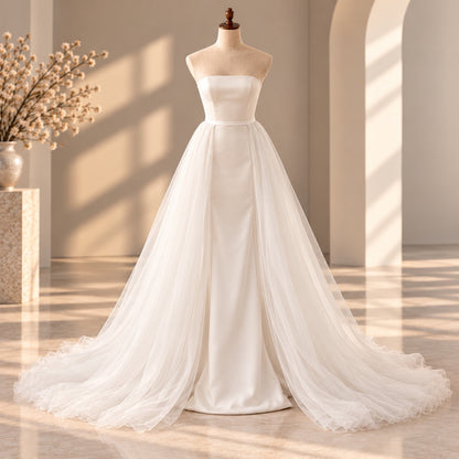 A strapless white wedding gown displayed on a mannequin in a softly lit interior with warm sunlight and shadows. The dress has a fitted bodice and a full layered tulle skirt with a long train.
