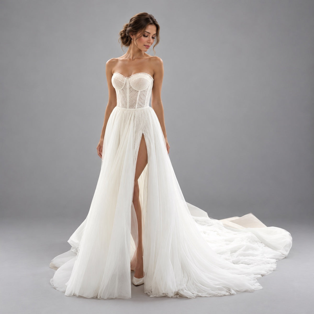 A woman modeling a strapless white wedding gown with a structured lace corset bodice, high front slit, and a long flowing tulle train against a plain gray studio background.