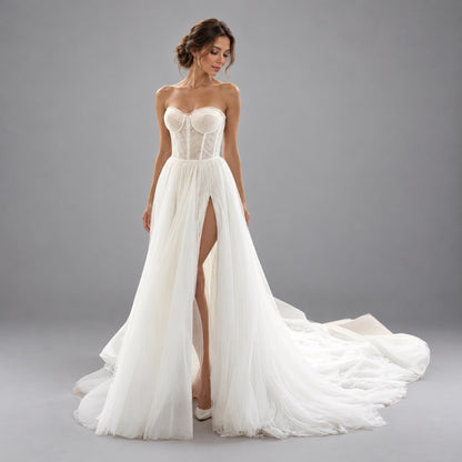 A woman modeling a strapless white wedding gown with a structured lace corset bodice, high front slit, and a long flowing tulle train against a plain gray studio background.