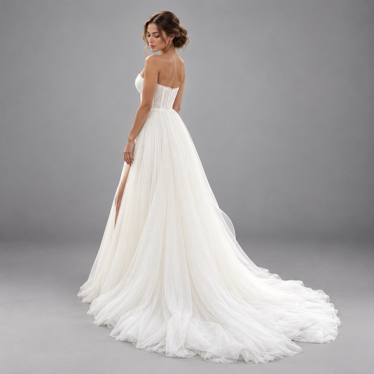 A woman modeling a strapless white wedding gown with a fitted lace bodice, sheer tulle skirt, and long train against a plain gray studio background.