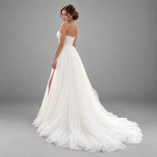 A woman modeling a strapless white wedding gown with a fitted lace bodice, sheer tulle skirt, and long train against a plain gray studio background.