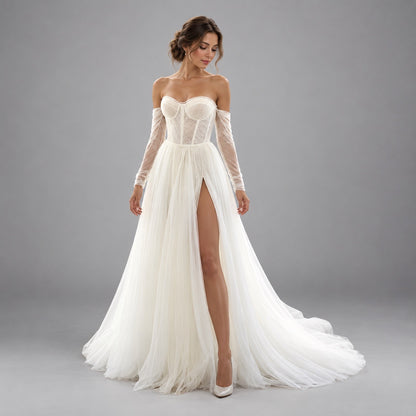A woman modeling an off-the-shoulder white wedding gown with a lace corset bodice, sheer long sleeves, a high thigh slit, and a flowing tulle skirt with a train, photographed against a plain gray studio background.