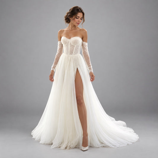A woman modeling an off-the-shoulder white wedding gown with a lace corset bodice, sheer long sleeves, a high thigh slit, and a flowing tulle skirt with a train, photographed against a plain gray studio background.